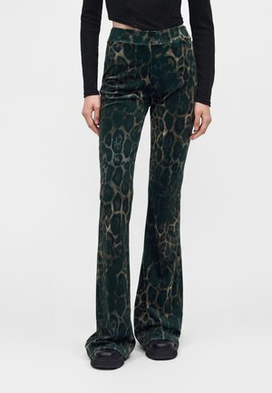Personne portant un pantalon évasé en velours imprimé léopard vert foncé, des chaussures plateformes noires épaisses et un haut court noir à manches longues.
