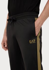 Black athletic pants with a relaxed fit, featuring yellow stripes along the side and an embroidered "EA7" logo on the front pocket.