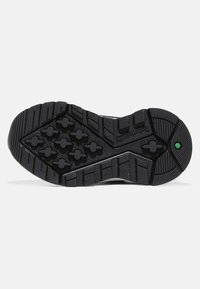 Suela negra de un zapato con una superficie texturizada, con surcos profundos y patrones hexagonales, y un pequeño detalle en verde.