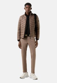 Lysebrun dunjakke med høj krave, sort skjorte under, beige bukser og hvide sneakers. Har en rygsæk på skulderen.