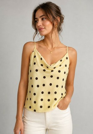 Femme aux cheveux bruns en chignon lâche, portant un caraco jaune à pois noirs et un pantalon blanc, main dans la poche, regardant sur le côté.