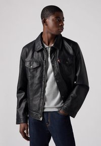 Veste en cuir noir avec un col, deux poches poitrine et fermeture à boutons. Portée par-dessus un pull gris et associée à un jean en denim foncé.