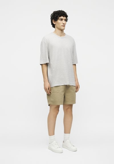 Νέος άνδρας στέκεται φορώντας μια γκρι και λευκή ριγέ φαρδιά μπλούζα, χακί σορτς, λευκές κάλτσες και λευκά αθλητικά σε απλό φόντο.