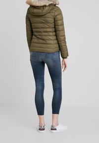 Chaqueta de plumas verde oliva con capucha de piel sintética desmontable, combinada con jeans skinny azul oscuro y zapatillas blancas con detalles plateados.