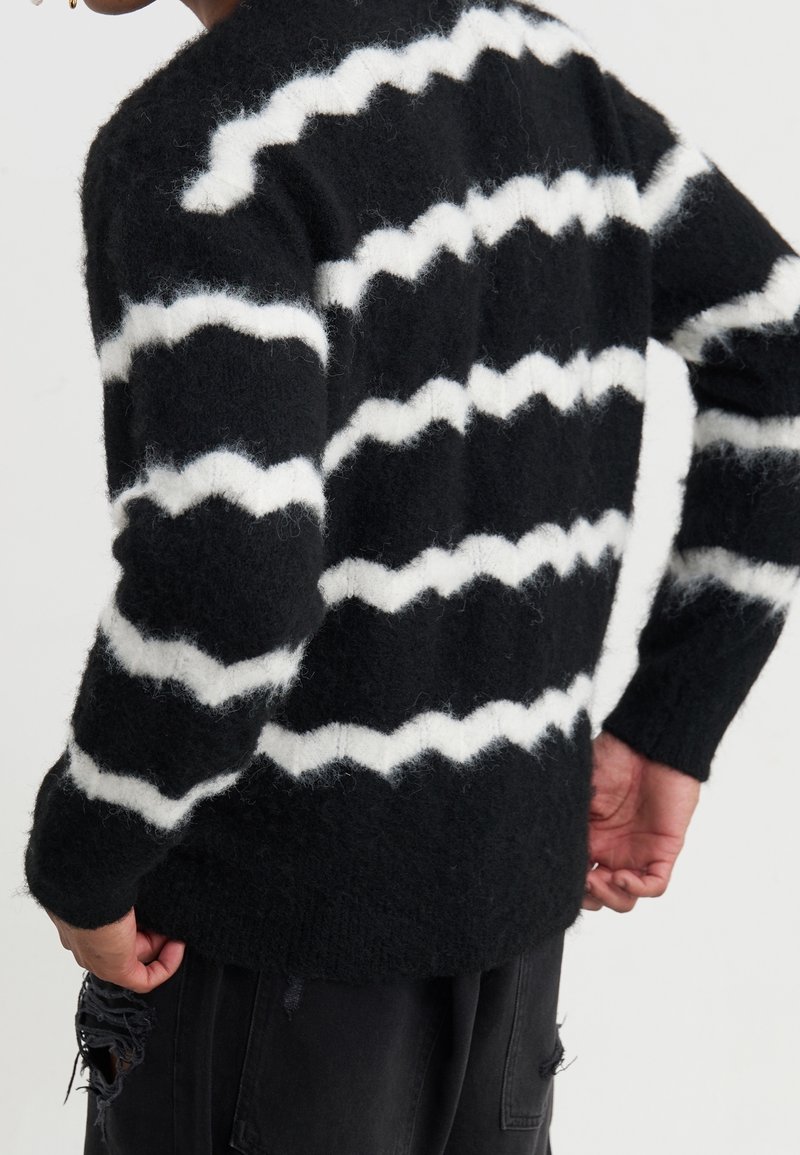 Personne portant un pull noir duveteux avec des rayures blanches en zigzag, associé à un pantalon noir déchiré, vue de dos.