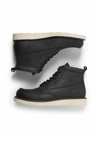 Botas de tobillo de cuero negro con suela de goma blanca, cordones en la parte delantera y un diseño cosido. Cuenta con una lengüeta en el talón para facilitar su uso.