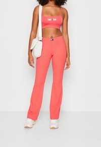 Kobieta w koralowym, dwuczęściowym zestawie z prążkowanej tkaniny z logo Tommy Jeans, białych sneakersach i białej torbie na ramię, stojąca na tle jednolitym.