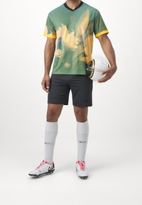 Grön och orange mönstrad Nike-tröja, svart krage; svarta shorts; vita Nike-skor med röda accenter; håller en vit och gul fotboll.