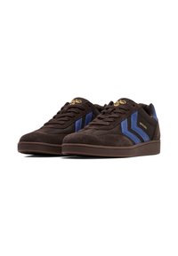 Brązowe zamszowe sneakersy z niebieskimi paskami i złotym logo na języku. Oferują gumową podeszwę oraz fakturowane akcenty z tkaniny.