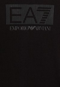 Logo EA7 s vertikálními pruhy nad textem Emporio Armani a emblémem orla na černém textilním pozadí.