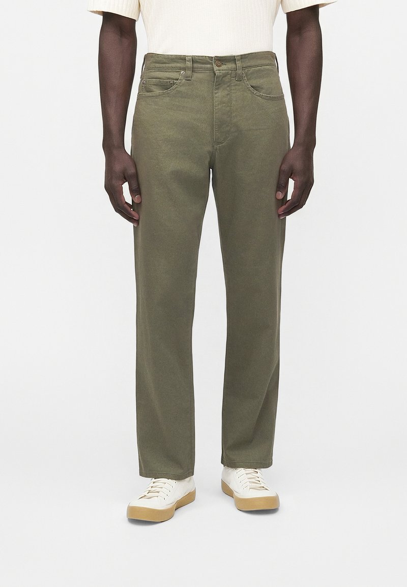 Άνδρας που φοράει ελαφρώς χακί παντελόνια με ίσια γραμμή, λευκά αθλητικά παπούτσια με καουτσούκ σόλες και ανοιχτόχρωμο κοντομάνικο μπλουζάκι, στέκεται πάνω σε λευκό φόντο.