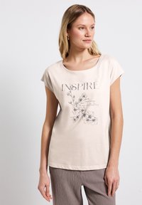 Camiseta de algodón de color beis claro, de manga corta. Presenta un gráfico floral y la palabra "INSPIRAR" en letras grandes. Textura suave.