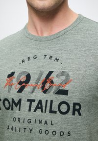 Γκρι μελανζέ T-shirt με στρογγυλή λαιμόκοψη, με μαύρη και πορτοκαλί γραφική εκτύπωση που περιλαμβάνει το "Tom Tailor" και "The Casual Brand 1962."