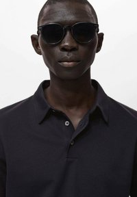 Svart polo skjorta med en texturerad tyg, två knappar vid kragen och smala ärmar. Modellen bär runda, mörka solglasögon.