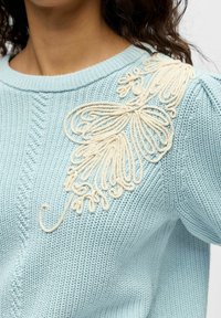 Pull bleu clair en maille avec un côtelé texturé et un motif floral brodé crème complexe sur l'épaule gauche.