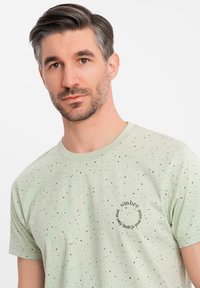 Camiseta verde claro con patrón salpicado negro. Presenta un gráfico redondo con el texto: "tu único límite es tu mente" en el frente.