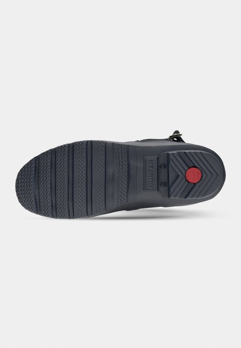 Suela de zapato de goma negra con textura, presenta un acento rojo en el talón. Diseño resistente con surcos pronunciados para mayor tracción.