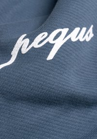 Sininen tekstuuripinta, jossa on kudottu kuvio, ja valkoista brodeerattua tekstiä, joka lukee "nequs." Materiaali vaikuttaa pehmeältä ja joustavalta.
