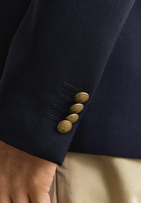 Mouw van een marineblazer met vier gouden decoratieve knopen met gegraveerde wapenschilden, gedragen over lichtbeige broek.