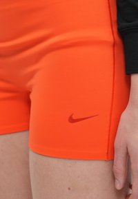 Calções atléticos laranja com uma textura suave apresentam um logo da Nike vermelho no canto inferior esquerdo e costuras planas para maior durabilidade.