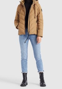 Personne portant une doudoune beige avec capuche, un col roulé noir, un jean bleu clair retroussé et des bottes de combat noires à lacets, debout sur un fond uni.