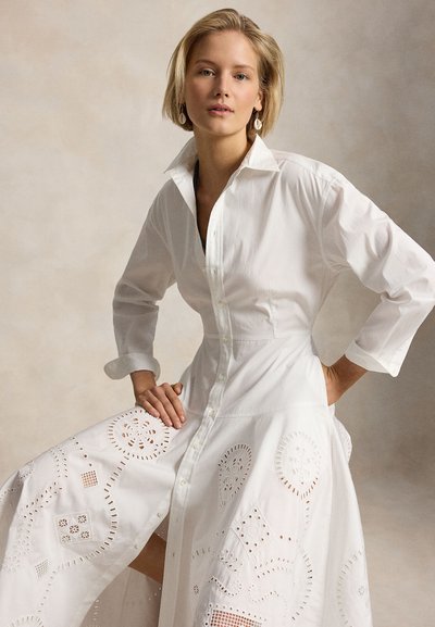 Femeie cu păr scurt și blond, îmbrăcată într-o rochie albă cu nasturi, având decupaje brodate, pozând cu o mână în talie și cealaltă ținând fusta.