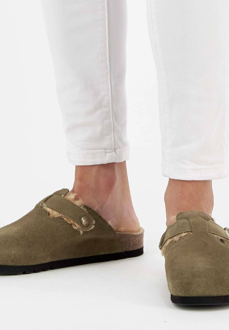 Sabots en daim verts avec une doublure en fourrure, design à enfiler, accent en matériel de bouton, dessus texturé, associés à un jean slim blanc.
