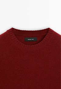 Burgundy stickad tröja med rundad halsringning. Tillverkad av mjukt material, har ribbad krage och märkesdetaljer vid halsen.
