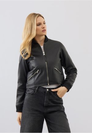 Blondynka w czarnej skórzanej kurtce bomber z zamykanymi kieszeniami i czarnych wysokich spodniach jeansowych, pozująca z jedną ręką w kieszeni.