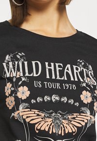 Czarny T-shirt z napisem "WILD HEARTS US TOUR 1978" w postarzanym białym kolorze, z kwiatowymi i motylkowymi grafikami w różowych i szarych akcentach.