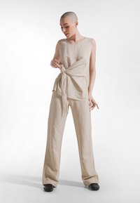 Beżowy lniany top bez rękawów z wiązaniem po boku, zestawiony z luźnymi beżowymi lnianymi spodniami. Prosta, gładka faktura o swobodnym kroju.