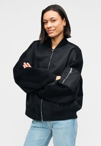 Μαύρο μπουφάν μπόμπερ με φερμουάρ μπροστά, ριμπ μανσέτες και συνδυασμό λείου και υφασμένου υφάσματος. Διαθέτει τσέπη με φερμουάρ σε γωνία στο μανίκι.