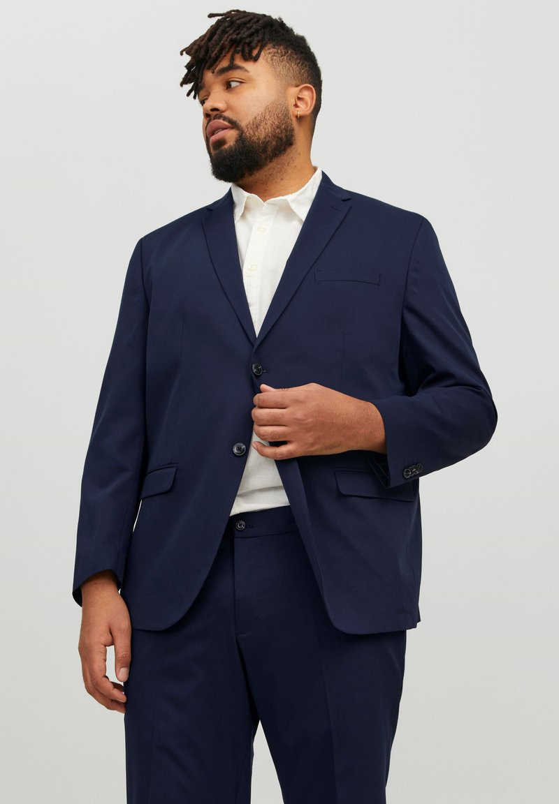 Marineblaues Sakko mit Revers, zwei Knöpfen und Fronttaschen. Kombiniert mit einem weißen Hemd. Glattes, leichtes Material.