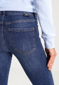 Jeans ajustados en denim azul oscuro con una textura suave, que cuentan con un bolsillo de parche y costuras amarillas. Llevados con un sudadera azul clara.