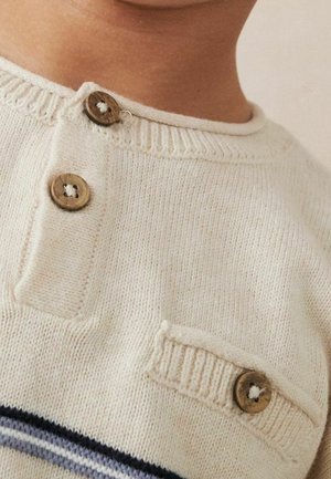 Pull en maille crème avec deux boutons en bois près de l'encolure et un sur une petite poche décorative.