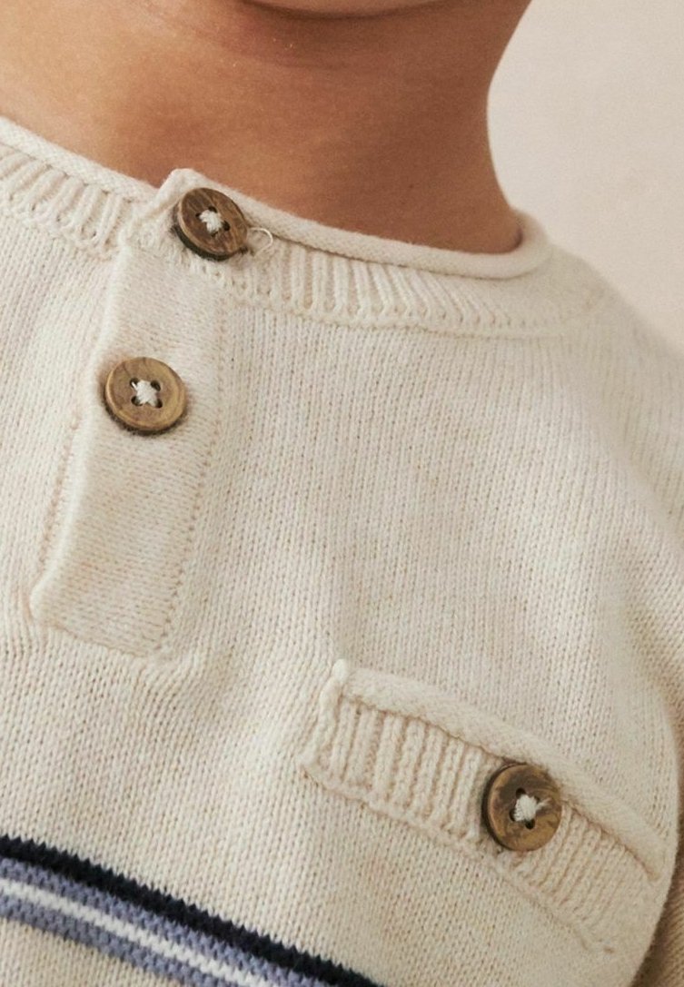 Pull en maille crème avec deux boutons en bois près de l'encolure et un sur une petite poche décorative.