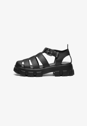 Black sandals made of leather, featuring multiple straps, an ankle buckle, and a chunky rubber sole with defined treads.