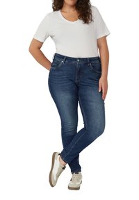 Donkerblauwe denim jeans met een slim fit, vijf-pocket ontwerp en lichte stiksel details, gecombineerd met een witte V-hals t-shirt en beige sneakers.