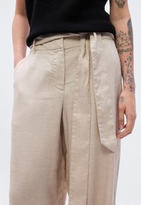 Μπεζ λινό παντελόνι με ψηλή μέση, με πιέτες, ζώνη δέσιμο και πλαϊνές τσέπες. Το ύφασμα έχει υφή.