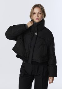 Veste courte matelassée noire avec col haut, tissu texturé, fermeture éclair et poches avant ; présente un ourlet côtelé et un détail de logo.