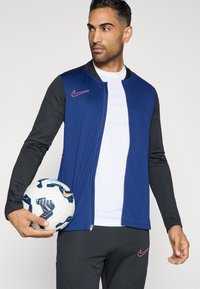 Μπλε και μαύρο αθλητικό μπουφάν με λεία υφή, κλείσιμο με φερμουάρ και ροζ λογότυπο Nike. Το μοντέλο κρατάει μία καλοσχεδιασμένη μπάλα ποδοσφαίρου.