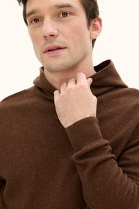 Brauner, gestrickter Pullover mit Kapuze, ausgestattet mit gerippten Bündchen und einer strukturierten Oberfläche. Eine Nahaufnahme zeigt eine Hand, die den Ausschnitt berührt.