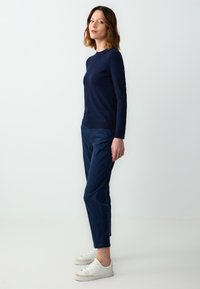 Vrouw met schouderlengte haar, gekleed in een marineblauwe trui, marineblauwe broek en witte sneakers, staande tegen een lichtgrijze achtergrond.
