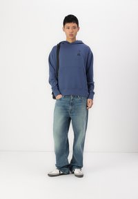 Μπλε φούτερ με μπροστινή τσέπη και κεντημένο λογότυπο, σε συνδυασμό με ανοιχτό μπλε φαρδύ τζιν και λευκά αθλητικά παπούτσια με γκρίζες λεπτομέρειες.