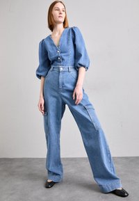 Denim jumpsuit med puffärmar, v-hals och framknäppning; ljusblå färg, vida ben och cargo-fickor på sidorna.