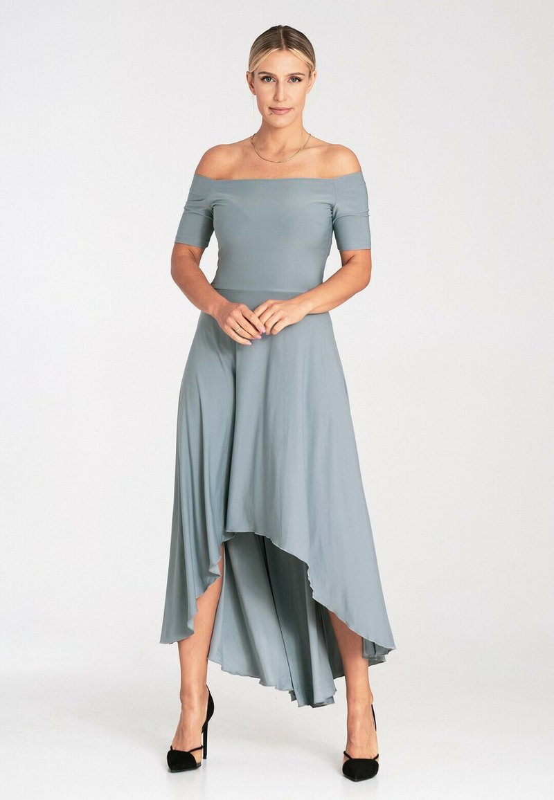 Sukienka w kolorze jasnoszarym, odsłaniająca ramiona, z asymetrycznym dołem i falbaną. Wykonana z gładkiego materiału, zestawiona z czarnymi szpilkami.