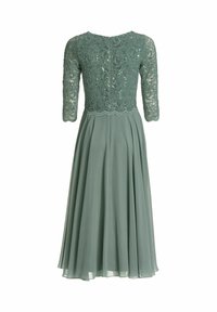 Vihreä pitsimekko, jossa on kolme neljänneksen mittaiset hihat. Tiukka yläosa, jossa on monimutkaiset pitsikoristeet, ja ilmava, laskostettu hame, joka on valmistettu kevyestä kankaasta.