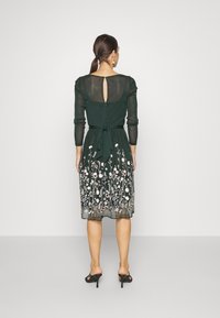 Dunkelgrünes Kleid mit langen, transparenten Ärmeln, floralem Stickereien am Rock und einem Schlüssellochdetail im Rücken. Mit gebundenem Taillengürtel und knielanger Länge.