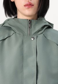 Hellgrüne Jacke mit durchgehendem Reißverschluss, Kapuze und Druckknopfverschluss. Das Material ist glatt und wirkt wasserabweisend, mit abgesteppten Nähten.