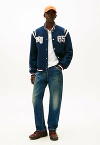 Chaqueta varsity azul con rayas blancas y rojas, número 85, llevada sobre una polo blanca. Combinada con jeans desgastados y zapatillas con acentos rojos. Gorra naranja.
